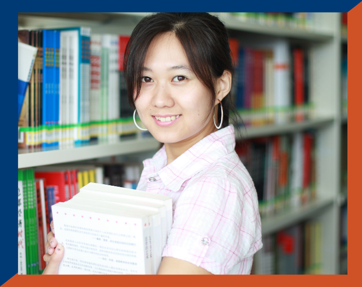 photo of woman with dark brown hair and wearing pink and white shirt smiling and holding stack of books.